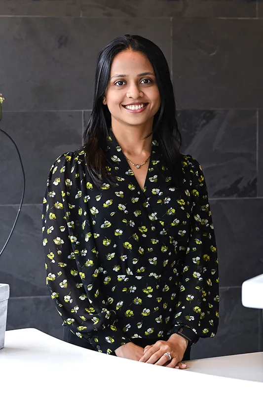 Maushami Patel, Patient Care Facilitators in a black and yellow patterned blouse standing in front of a dark wall
