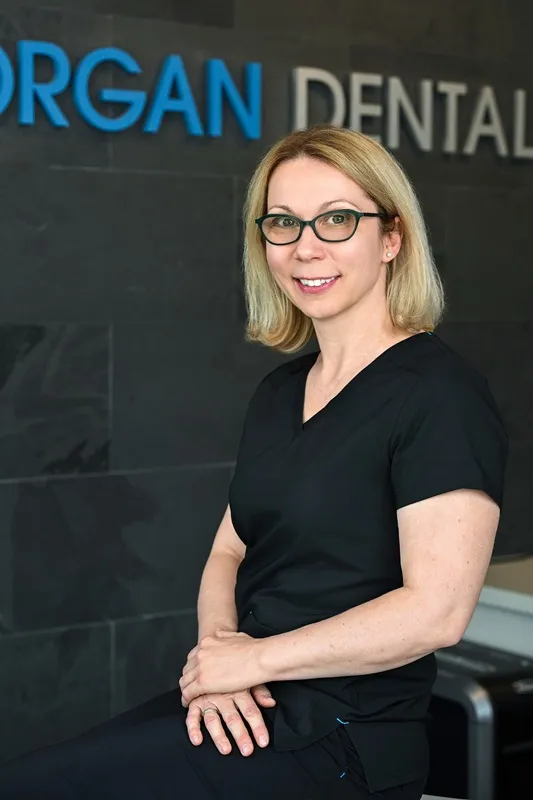 Olesia Borets, Registered Dental Hygienist with glasses wearing dark scrubs and sitting in front of a Morgan Dental sign