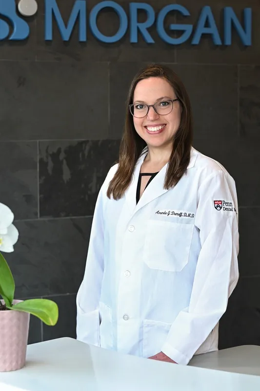 Dr. Amanda Dumoff wearing a white lab coat and smiling in front of a Morgan Dental logo